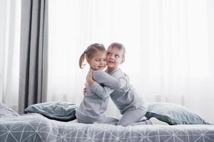 enfants heureux jouant dans la chambre blanche. petit garçon et fille, frère et soeur jouent sur le lit en pyjama. intérieur de pépinière pour enfants. vêtements de nuit et literie pour bébé et tout-petit. famille à la maison photo