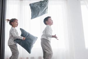 enfants jouant dans le lit des parents. les enfants se réveillent dans une chambre blanche ensoleillée. garçon et fille jouent en pyjama assorti. vêtements de nuit et literie pour enfant et bébé. intérieur de pépinière pour enfant en bas âge. matin en famille photo