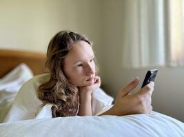 une fille dans une blanc chemise une adolescent mensonges sur une blanc lit dans sa mains elle détient une noir iphone 13 elle regards à le écran calmement élevage sa mains en haut photo