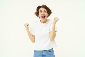 excité brunette fille, étudiant gagnant prix, célébrer la victoire, triomphant, permanent dans blanc T-shirt et jeans plus de blanc Contexte photo