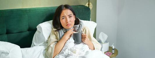 santé, grippe et gens concept. portrait de coréen femme sentiment malade, capture une froid, rester dans lit indisposé, en buvant chaud thé photo