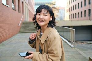 Jeune asiatique femme écoute la musique dans sans fil écouteurs, les usages téléphone intelligent sur rue, des promenades autour ville sur ensoleillé jour, souriant Heureusement photo