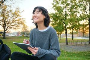 Jeune coréen femme est assis dans parc, dessine sur numérique tablette, écrit dans gadget avec stylo outil, regards autour et fait du Remarques, crée ouvrages d'art ou les taches photo