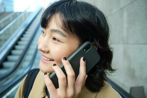 photo du visage de souriant coréen femme avec téléphone intelligent, fait du une téléphone appel, va vers le bas escalier mécanique dans ville, les déplacements à Université