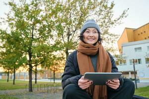 portrait de asiatique fille dans chaud vêtements, est assis sur banc avec numérique tablette et graphique stylo, souriant Heureusement, dessine en plein air dans froid temps photo