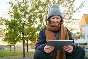 portrait de asiatique fille dans chaud vêtements, est assis sur banc avec numérique tablette et graphique stylo, souriant Heureusement, dessine en plein air dans froid temps photo