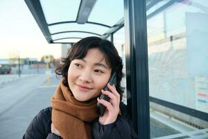 proche en haut portrait de mignonne Jeune femme appel quelqu'un, attendre sur autobus Arrêtez avec téléphone intelligent, en utilisant Téléphone tandis que attendant Publique transport à arrivée photo