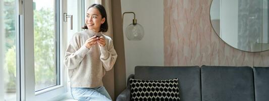 portrait de Jeune asiatique femme séance près fenêtre et à la recherche dehors, en buvant chaud café de Expresso tasse et profiter sa confortable journée de à Accueil photo