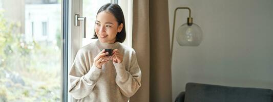 portrait de magnifique asiatique femme séance à Accueil avec tasse de café, profiter sa Expresso tandis que à la recherche à l'extérieur fenêtre à passant, souriant Heureusement photo