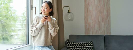 portrait de magnifique asiatique femme séance à Accueil avec tasse de café, profiter sa Expresso tandis que à la recherche à l'extérieur fenêtre à passant, souriant Heureusement photo