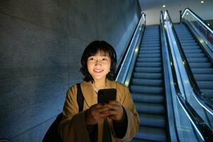 mignonne Jeune femme Aller vers le bas le escalier mécanique à le tube, en utilisant métro métro à commuer à travail ou université, permanent avec téléphone intelligent photo