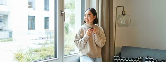 magnifique Jeune asiatique femme séance près fenêtre et en buvant sa café, en portant Expresso tasse et à la recherche à l'extérieur avec détendu, souriant visage expression photo