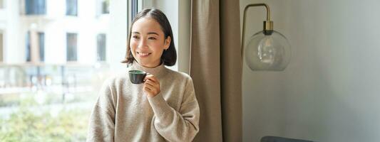 portrait de magnifique asiatique femme séance à Accueil avec tasse de café, profiter sa Expresso tandis que à la recherche à l'extérieur fenêtre à passant, souriant Heureusement photo