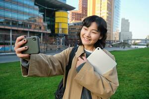 content Université étudiant, coréen fille prend selfie avec sa papiers et numérique tablette, détient téléphone intelligent et pose près Université Campus photo