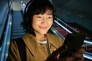 proche en haut de mignonne asiatique fille dans écouteurs, choix chanson à Ecoutez tandis que faire la navette, Aller vers le bas escalier mécanique à métro, souriant comme en train de lire message sur mobile téléphone photo