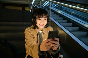 technologie. portrait de asiatique fille est assis sur escaliers près escalier mécanique, écoute la musique dans écouteurs et les usages social médias, lit, montres vidéo dans Publique endroit photo