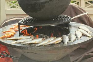 grillé poisson sur une en forme de bol plat gril avec Feu trou dans le centre. Extérieur Fruit de mer barbecue cuisson. pique-nique sur arrière-cour sur ouvert air. rue nourriture concept. en train de préparer poisson plus de ouvert feu de camp photo