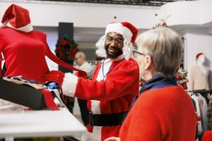 boutique employé guidage client dans boutique, portant de fête Père Noël claus costume tandis que portion les clients avec Noël présent des idées et suggestions. Jeune homme recommander chemisier à Sénior femme. photo