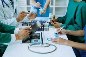 médical équipe ayant une réunion avec médecins dans blanc laboratoire manteaux et chirurgical gommages assise à une table discuter une les patients travail en ligne en utilisant des ordinateurs dans le médical industrie photo