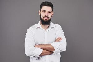 jeune bel homme appuyé contre un mur gris avec les bras croisés. un jeune homme sérieux avec une barbe regarde la caméra photo