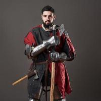 chevalier médiéval sur fond gris. portrait d'un guerrier brutal au visage sale avec une armure de cotte de mailles, des vêtements rouges et noirs et une hache de combat photo