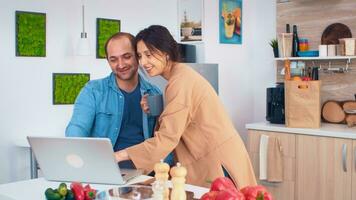 femme à la recherche à mari portable tandis que il est travail dans cuisine. petite amie en portant tasse de café. mari et épouse cuisine recette aliments. content en bonne santé ensemble mode de vie. famille recherche pour en ligne repas. santé Frais salade photo