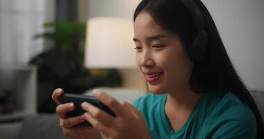 portrait de Jeune asiatique femme portant des lunettes et écouteurs jouit en jouant en ligne esport Jeux sur téléphone intelligent séance sur canapé dans le vivant pièce à maison, joueur mode de vie concept. photo