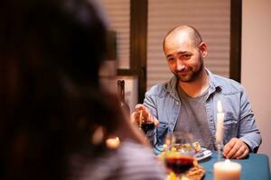 homme à la recherche à épouse tandis que ayant romantique dîner et en portant verre avec rouge du vin. parlant content séance à table à manger chambre, profiter le repas à Accueil ayant romantique temps à bougie lumières. photo