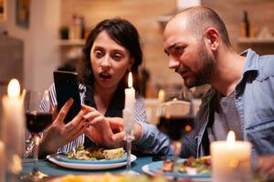 mari et épouse à la recherche sous le choc à téléphone tandis que ayant romantique dîner dans cuisine adultes séance à le table dans le cuisine navigation, recherche, en utilisant smartphone, l'Internet, célébrer anniversaire. photo