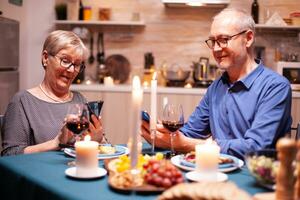 retraité homme souriant en utilisant téléphone et ayant une conversation avec épouse dans cuisine dans temps de dîner. séance à le table dans le à manger chambre, navigation, recherche, en utilisant téléphone, l'Internet, photo