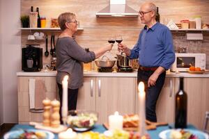 Sénior couple célébrer anniversaire avec rouge du vin dans à manger chambre. vieilli couple dans l'amour parlant ayant agréable conversation pendant en bonne santé repas. photo