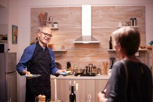 vieux homme parlant avec épouse tandis que portion sa dîner pendant relation fête. Sénior couple en parlant, séance à le table dans cuisine, profiter le repas, célébrer leur anniversaire dans le à manger chambre. photo