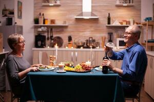 personnes âgées homme ouverture une bouteille de du vin dans cuisine pendant romantique dîner avec épouse . Sénior vieux couple en parlant, séance à le table dans à manger pièce , profiter le repas, célébrer leur anniversaire. photo