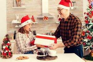 souriant nièce célébrer Noël recevoir présent boîte avec rouge arc de grand-père. Sénior homme portant Père Noël chapeau surprenant petite fille avec hiver vacances cadeau dans Accueil cuisine avec Noël arbre dans le Contexte. photo