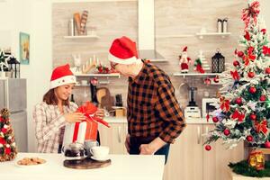 souriant nièce vérification Noël cadeau boîte avec rouge arc de personnes âgées homme dans cuisine. Sénior homme portant Père Noël chapeau surprenant petite fille avec hiver vacances présent dans cuisine avec Noël arbre dans le Contexte. photo