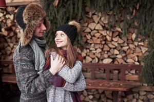 concept d'hiver, de vacances, de couple, de noël et de personnes - homme et femme souriants en chapeaux et écharpe étreignant sur une maison de campagne en bois et un fond de neige