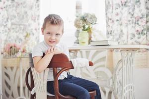 petit garçon en jeans pantalon bleu est assis sur la chaise en bois dans la pièce. fait des devoirs. retour à l'école photo
