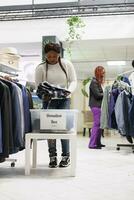 africain américain client faire un don vêtements à aide sociale organisation dans achats centre. femme en mettant Nouveau et seconde main vêtements dans don boîte tandis que achats dans branché boutique photo
