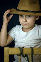 une concis portrait de une européen garçon. portrait sur une foncé Contexte de une garçon dans une cow-boy chapeau et une blanc T-shirt. photo