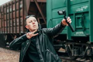 Beau gars dans noir vêtements et des lunettes de soleil fabrication selfie ou diffusion vidéo en utilisant action caméra avec cardan caméra stabilisateur à chemin de fer. homme fait du photo contre le cargaison wagons