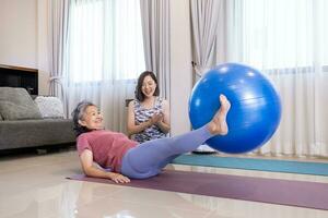 Sénior asiatique femme est exercice à Accueil en utilisant yoga Balle pour coeur corps et abdomen muscle bâtiment avec sa entraîneur fille encourager et applaudissement pour aîné en bonne santé concept photo