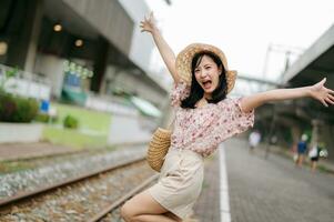 Jeune asiatique femme voyageur avec tissage panier content souriant à la recherche à une caméra à côté de train chemin de fer. périple voyage mode de vie, monde Voyage explorateur ou Asie été tourisme concept. photo