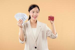 portrait de content réussi sur de soi Jeune asiatique affaires femme portant blanc veste en portant en espèces argent dollars et crédit carte permanent plus de beige Contexte. millionnaire entreprise, achats concept photo