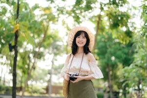 portrait de asiatique Jeune femme voyageur avec tissage chapeau et panier et une caméra sur vert Publique parc la nature Contexte. périple voyage mode de vie, monde Voyage explorateur ou Asie été tourisme concept. photo