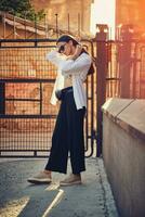 portrait de une fille dans foncé des lunettes de soleil posant dans ville. habillé dans Haut avec floral imprimer, blanc chemise, noir pantalon, taille sac et baskets. photo