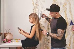 mignonne peu fille et sa tatoué papa sont en jouant ensemble près une miroir. papa est Faire le sien fille cheveux. famille vacances et unité. photo