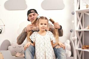 marrant temps tatoué père dans une casquette et le sien enfant sont en jouant à maison. papa est Faire le sien fille cheveux dans sa chambre à coucher. famille vacances et unité photo