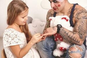 marrant temps tatoué père dans une casquette et le sien enfant sont en jouant à maison. mignonne fille est Faire maquillage à sa papa dans sa chambre à coucher. famille vacances unité photo