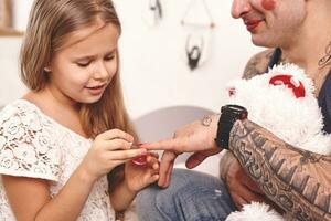 marrant temps tatoué père dans une casquette et le sien enfant sont en jouant à maison. mignonne fille est Faire maquillage à sa papa dans sa chambre à coucher. famille vacances unité photo