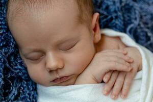 nouveau née - bébé, visage fermer. le en train de dormir nouveau née garçon en dessous de une blanc tricoté couverture mensonges sur le bleu fourrure. photo